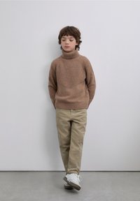 Suéter marrón de cuello alto tejido con textura acanalada, combinado con pantalones beige y zapatillas deportivas blancas, sobre un fondo liso.