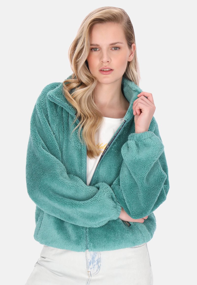 Blaugrüne flauschige Jacke mit hohem Kragen, Reißverschluss und weiten Ärmeln. Strukturierter Stoff mit weichem Aussehen, getragen über einem weißen Hemd.