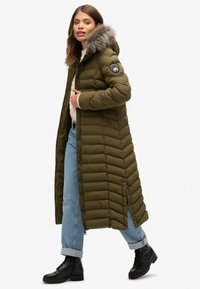Lange olijfgroene doffe pufferjas met een met bont gevoerde capuchon, zijzakken met rits en chevronstiksel. Gestyled met opgerolde jeans en zwarte laarzen.