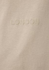 Tessuto di cotone beige con lettering in rilievo "LONDON" in una tonalità sottile e coordinata, che crea un effetto di texture sulla superficie liscia.