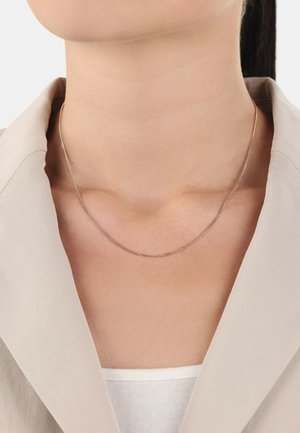 Collana in argento con un design delicato e sottile, che poggia sul décolleté, abbinata a un blazer beige chiaro e a una maglietta bianca.