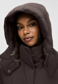 Doudoune marron avec un design matelassé et une grande capuche. Dotée de boutons-pression et d'une doublure intérieure brillante, elle évoque chaleur et protection.