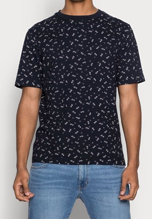 Mand iført en navyblå kortærmet t-shirt med små hvide geometriske former, kombineret med lyseblå jeans, stående mod en ensfarvet baggrund.