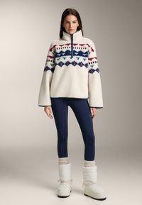 Fleecepullover i krämfärg med halvlång dragkedja och flerfärgade geometriska mönster på bröstet. Kombinerad med marinblå leggings och vita stövlar.
