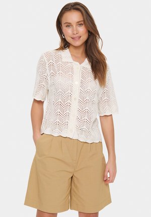 Blusa bianca in crochet con orlo a zigzag e maniche corte, caratterizzata da una chiusura con bottoni e un motivo a zigzag, abbinata a pantaloni corti beige.