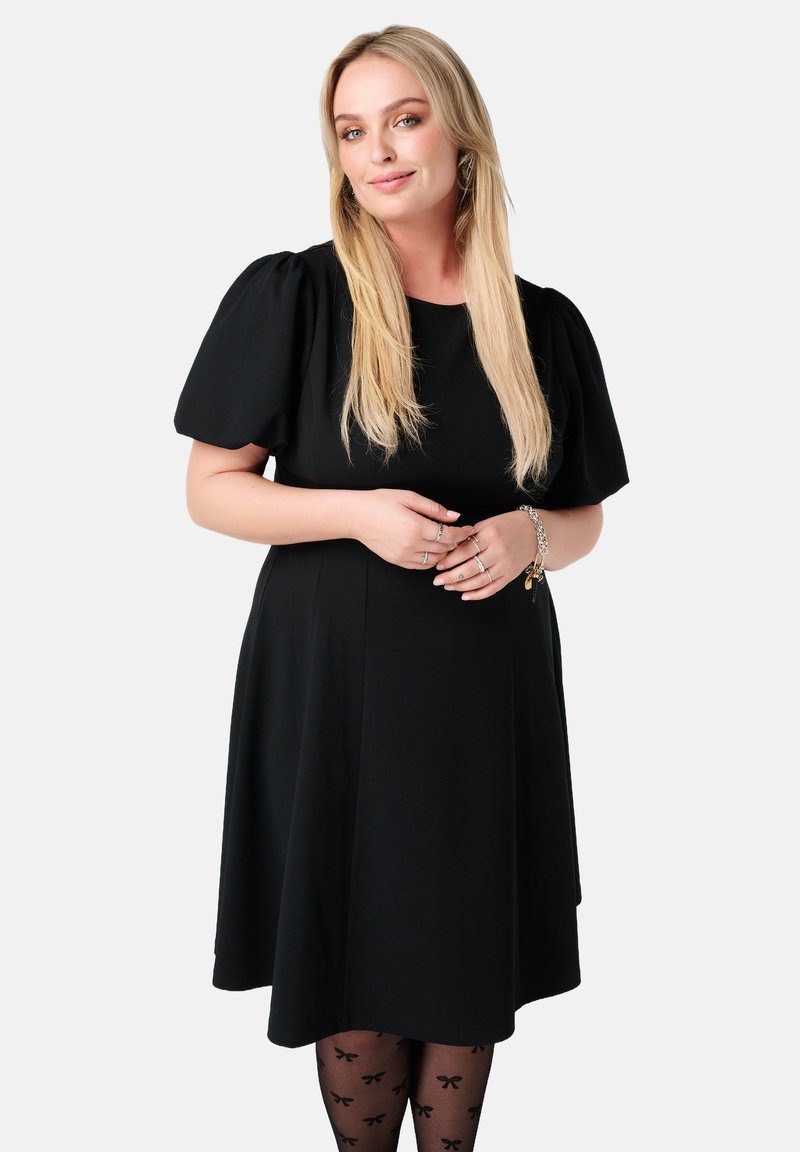 Zwarte jurk met korte, pofmouwen, een aansluitend lijfje en een A-lijn silhouette. Model draagt zwarte panty’s met strikpatronen en zilveren sieraden.