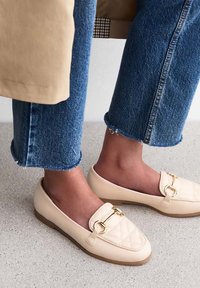 Beiga vadderade loafers med ett guldtonat spänne, i kombination med avklippta blå jeans med fransiga nederkanter, på en ljus texturerad yta.