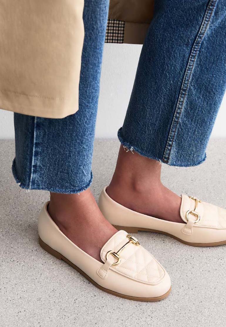 Beiga vadderade loafers med ett guldtonat spänne, i kombination med avklippta blå jeans med fransiga nederkanter, på en ljus texturerad yta.