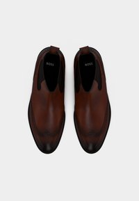 Chaussures en cuir marron au design élégant, présentant un bout texturé et des coutures décoratives le long des bords. Modèle à enfiler avec des côtés élastiques.