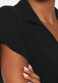 Vestito nero con colletto e maniche corte. Tessuto liscio con silhouette aderente. Tono della pelle visibile sotto la manica.