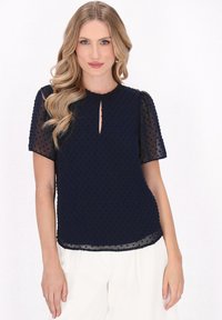 Marineblå bluse med prikket tekstur, korte gennemsigtige ærmer og nøglehul-udskæring. Flydende design med en let buet søm.