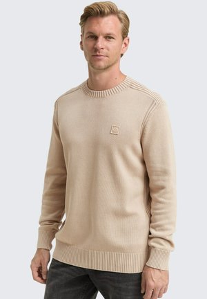 Blonder Mann, der einen beigen Strickpullover mit gerippten Ärmeln und Saum trägt, steht vor einem schlichten, hellen Hintergrund und schaut in die Kamera.