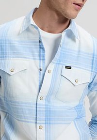 Ανοιχτό μπλε πουκάμισο με καρό σχέδιο, κουμπώνει με κουμπιά, έχει δύο μπροστινές τσέπες με καπάκια και χρυσούς κουμπιά. Είναι ορατή η απαλότητα του υφάσματος.