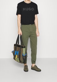 Camiseta de algodón negra con estampado "HUGO", pantalones verdes ajustados, zapatillas de olivo, sosteniendo una bolsa de tote con múltiples bolsillos en verde, negro y azul.