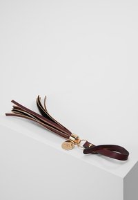 Porte-clés à gland en cuir avec lanières marron foncé, anneau doré et petit charm rond doré, posé sur une surface blanche.