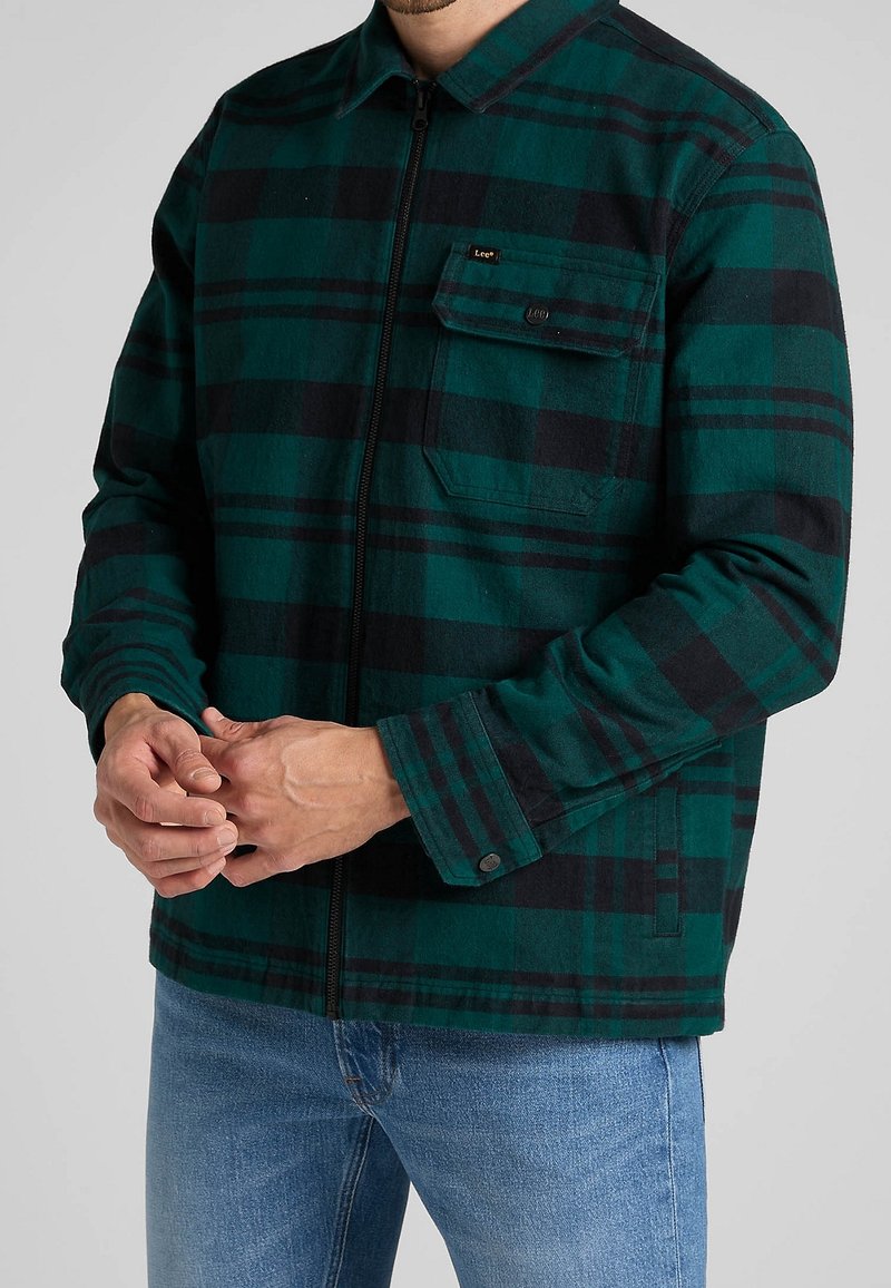 Homme portant une veste zippée à carreaux verts et noirs avec une poche poitrine et un jean bleu clair, mains jointes devant.
