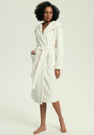 White ribbed bathrobe with a hood, waist tie, and two front pockets. Soft texture. Model standing barefoot against a neutral background.