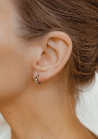 Orecchino a cerchio in argento con design a catena attorcigliata, caratterizzato da una finitura liscia. Indossato sul lobo dell'orecchio contro i capelli castano chiaro.