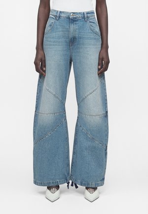 Modelka má na sebe svetlomodré široké rifle so švami patchwork a biele lodičky so špicatou špičkou na bielom pozadí.