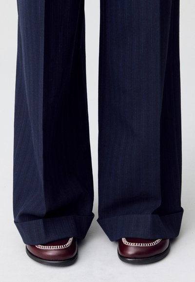 Pantaloni a gamba larga navy con righe, con orli risvoltati, abbinati a scarpe in pelle bordeaux con dettagli di cucitura bianca.