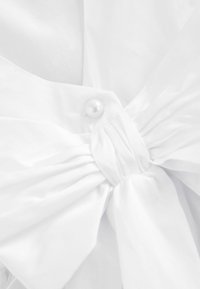 Tissu blanc avec une texture douce et lisse, présentant un motif noué et un bouton en perle décoratif. Détail simple et élégant.