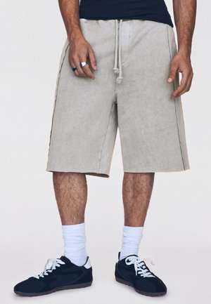 Mann trägt beige knielange Shorts mit Kordelzug, weiße Socken, schwarze Sneakers mit weißen Schnürsenkeln und Ringe an den Fingern der linken Hand.