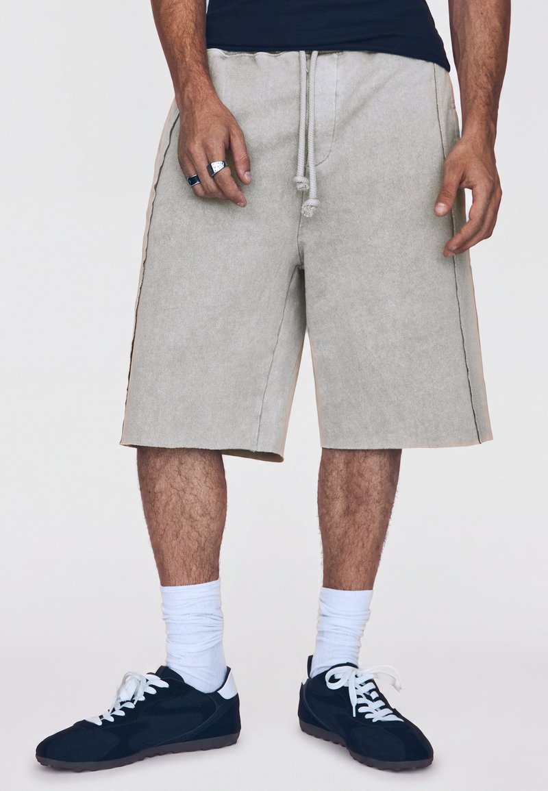 Homme portant un short beige mi-long à la taille avec un cordon, des chaussettes blanches, des baskets noires avec des lacets blancs, et des bagues aux doigts de la main gauche.