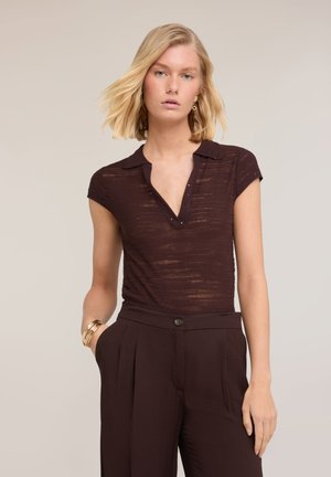 Blondine met kort haar die een donkerbruine doorschijnende top met korte mouwen en kraag draagt en een donkerbruine broek met hoge taille.