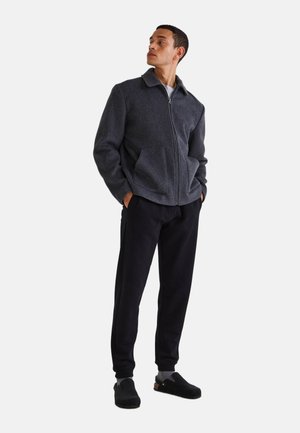 Veste zippée gris foncé avec un col, deux poches avant et un tissu texturé, associée à un pantalon noir et des chaussures noires.