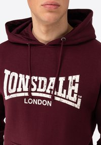 Bordeaux hoodie met een kanga zak, voorzien van een groot versleten wit "LONSDALE LONDON" logo bedrukt op de voorkant en met trekkoorden aan de capuchon.