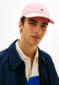 Ροζ βαμβακερό καπέλο baseball με μικρό πολύχρωμο λογότυπο. Φορεμένο με μπλε σκούρο μπουφάν και πουκάμισο με γιακά με μπλε και λευκές λεπτομέρειες.