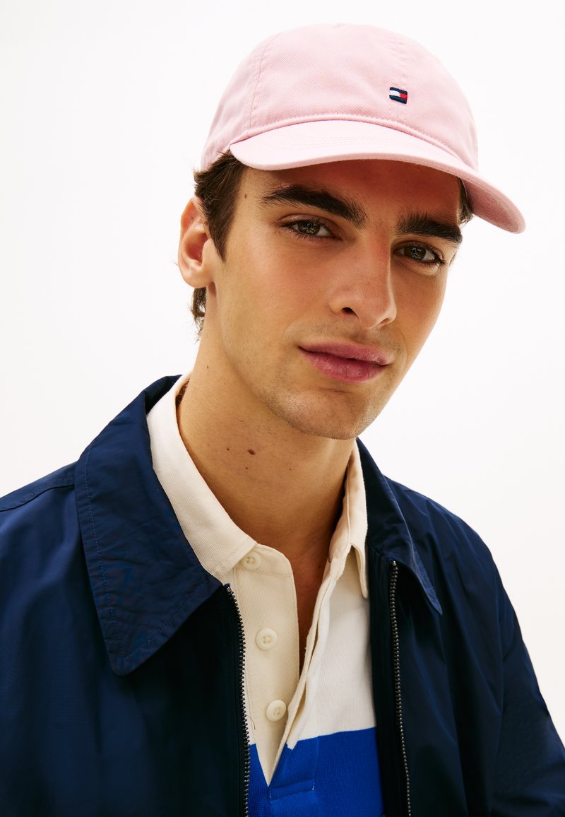 Ροζ βαμβακερό καπέλο baseball με μικρό πολύχρωμο λογότυπο. Φορεμένο με μπλε σκούρο μπουφάν και πουκάμισο με γιακά με μπλε και λευκές λεπτομέρειες.