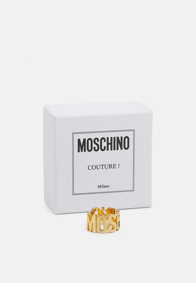 Gouden ring met het opschrift "MOSCHINO", met een uitgesneden ontwerp. Begeleidt door een witte textuurdoos met het label "MOSCHINO COUTURE!"