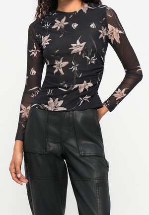Femme portant un haut en mesh noir à manches longues avec un imprimé floral rose et un pantalon en cuir noir taille haute avec de grandes poches.