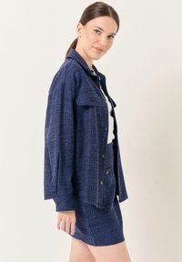Giacca tessuta blu navy con grandi bottoni frontali e finitura testurizzata, abbinata a una gonna coordinata. Entrambi hanno una vestibilità comoda.