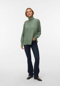Svijetlozeleni turtleneck pulover od mekanog materijala, labavog kroja s bočnim prorezima, uparen s tamnoplavim trapericama u zvonastom kroju i crnim balerinkama.