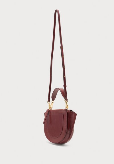 Δερμάτινη τσάντα crossbody σε μπορντό χρώμα με καμπυλωτό καπάκι, ρυθμιζόμενο λουρί, μικρή λαβή και κούμπωμα σε χρυσό τόνο σε ανοιχτό φόντο.