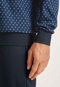 Marineblauwe sweatshirt met een klein bloemmotief in lichtblauw, met ribgebreide manchetten. Hand rustend op een donker gekleurde stof eronder.