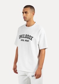 Weißes Oversize-T-Shirt mit schwarzem "SMILODOX ATHL. WEAR" Text, Rundhalsausschnitt und kurzen Ärmeln, kombiniert mit weißen Jogginghosen.