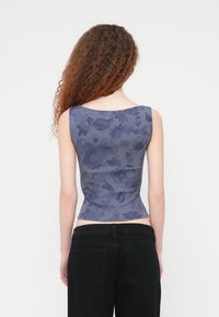 Αμάνικο εφαρμοστό τοπ σε σκούρο μπλε με floral σχέδιο, υφασμάτινο με υφή. Φορεμένο με μαύρα τζιν, παρουσιάζοντας καμπύλο σχέδιο στην πλάτη.