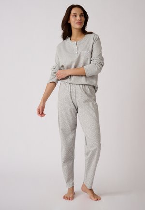 Mujer de pie descalza, llevando una camiseta de manga larga gris claro y pantalones grises a juego con pequeños lunares blancos sobre un fondo liso.