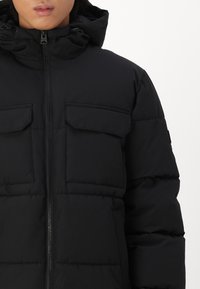 Svart puffjakke med hette, frontreisesykkel og to klafflommer; laget av glatt, isolert materiale med en strukturert design.