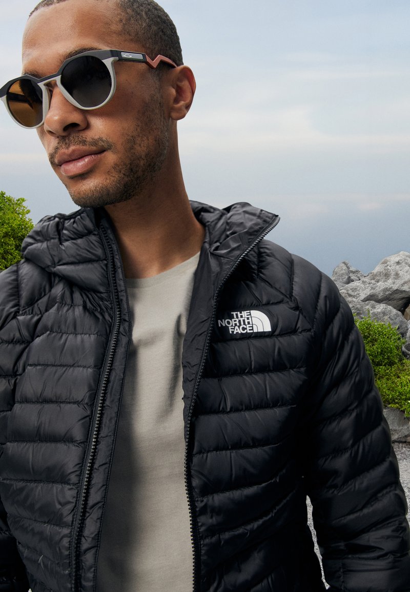 Zwarte pufferjack met capuchon, voorzien van een horizontaal doorgestikt patroon en het logo van The North Face. De persoon draagt ronde zonnebrillen met een grijs en zwart frame.