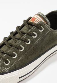 Baskets basses Converse en daim vert olive avec lacets noirs, semelle en caoutchouc blanche, œillets métalliques et étiquette de languette marquée.