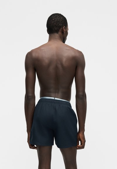 Άνδρας που στέκεται με την πλάτη, χωρίς μπλούζα, φοράει σκούρα μπλε μαγιό με ανοιχτό μπλε ζώνη μέσης, μπροστά σε ένα απλό ανοιχτό φόντο.