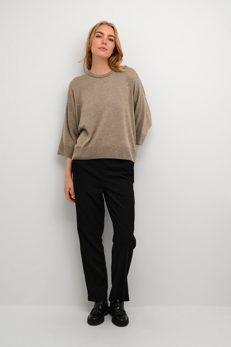 Beige, oversize pletený svetr s kulatým výstřihem, spárovaný s černými kalhotami na míru a černými botami, vše na neutrálním pozadí.