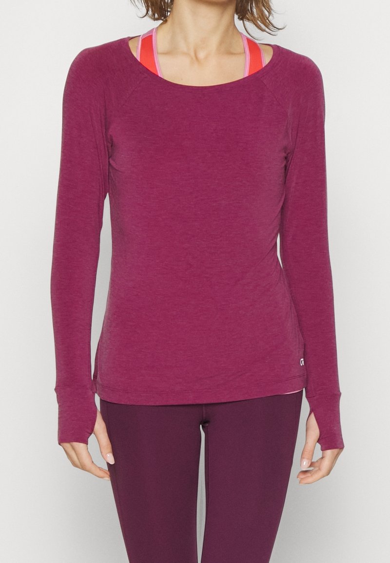 Sportief shirt met lange mouwen in kastanjebruine stof met een zachte textuur, voorzien van een brede halslijn en manchetten bij de polsen, gedragen over een roze racerback.