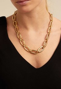 Collar de cadena de oro compuesto por grandes eslabones ovalados con un acabado pulido, llevado sobre una blusa negra con un diseño de escote en V.