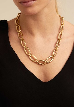 Collier en or composé de grands maillons ovales avec une finition polie, porté sur un haut noir à décolleté en V.