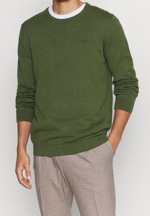 Mand iført en olivengrøn langærmet sweater over en hvid undertrøje og lysebrune ternede bukser med den ene hånd i lommen.